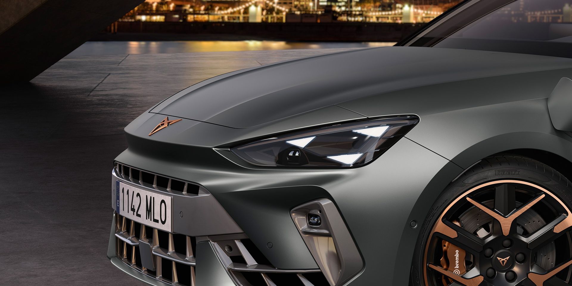 Detailný pohľad na prednú časť šedého športového vozidla CUPRA s moderným dizajnom. V pozadí sú osvetlené budovy mesta za súmraku. Auto má koleso s čiernym diskom s bronzovými prvkami.