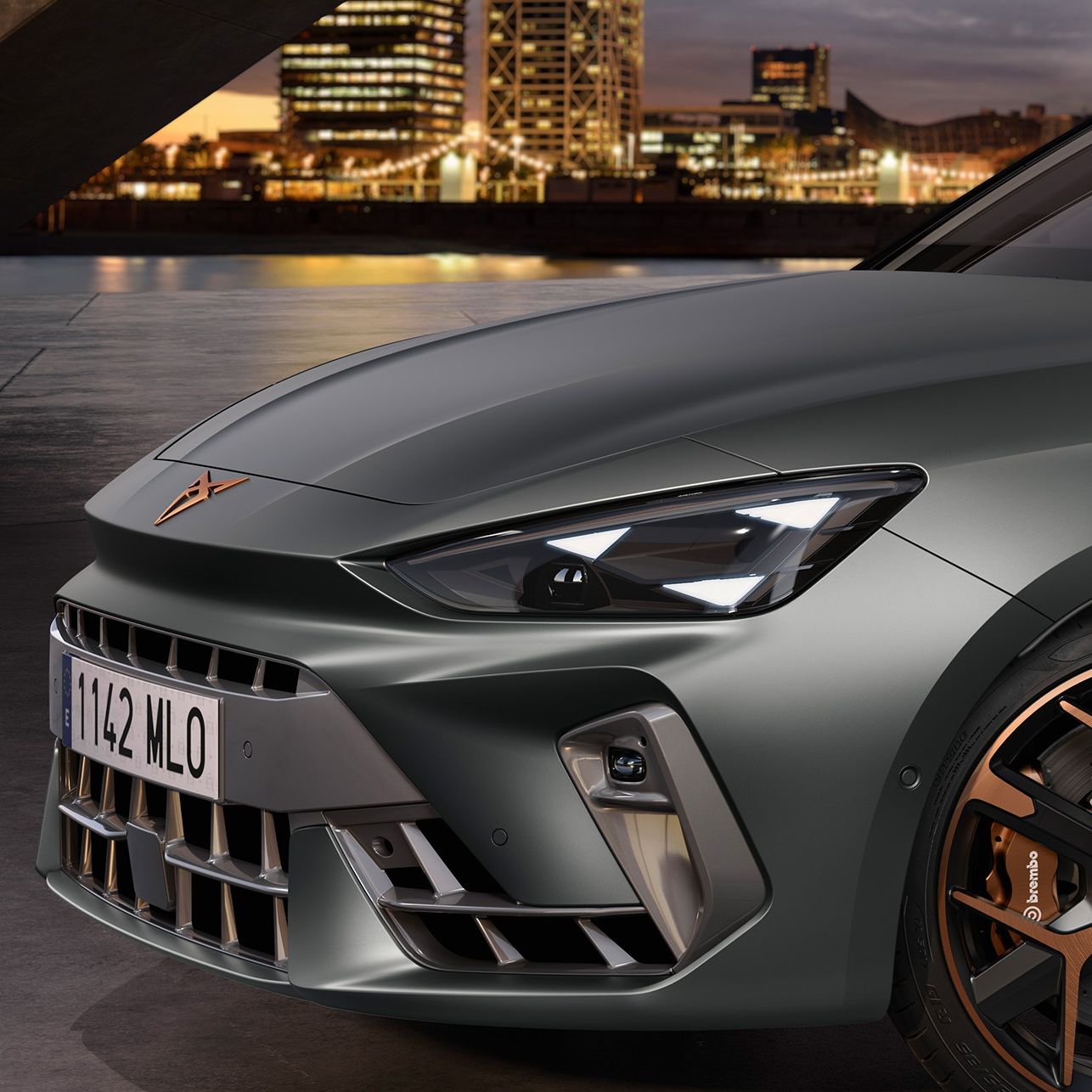 Detailný pohľad na prednú časť šedého športového vozidla CUPRA s moderným dizajnom. V pozadí sú osvetlené budovy mesta za súmraku. Auto má koleso s čiernym diskom s bronzovými prvkami.