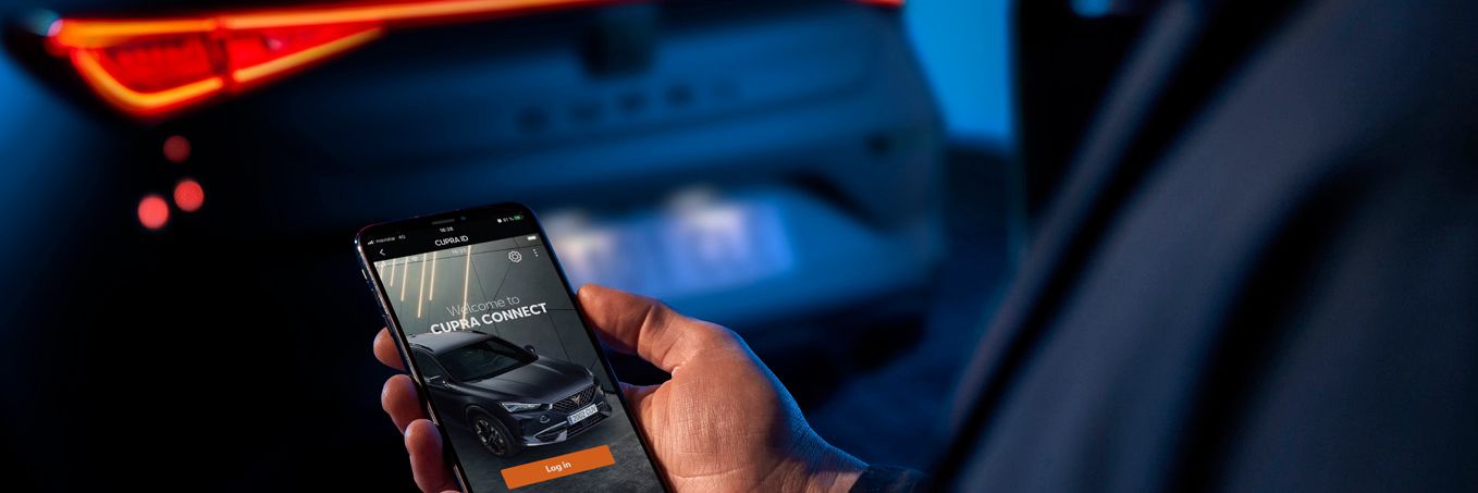 Osoba drží mobilný telefón, na ktorom je zobrazená aplikácia na ovládanie vozidla. V pozadí je vidieť zadnú časť tmavého auta s rozsvietenými svetlami. Prostredie je osvetlené modrým svetlom.