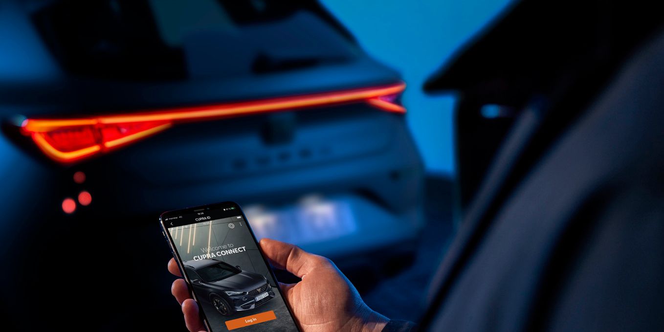 Osoba drží mobilný telefón, na ktorom je zobrazená aplikácia na ovládanie vozidla. V pozadí je vidieť zadnú časť tmavého auta s rozsvietenými svetlami. Prostredie je osvetlené modrým svetlom.