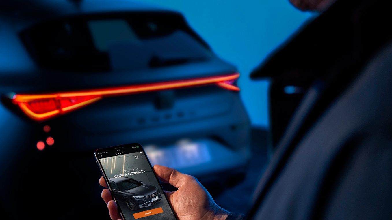 Osoba drží mobilný telefón, na ktorom je zobrazená aplikácia na ovládanie vozidla. V pozadí je vidieť zadnú časť tmavého auta s rozsvietenými svetlami. Prostredie je osvetlené modrým svetlom.