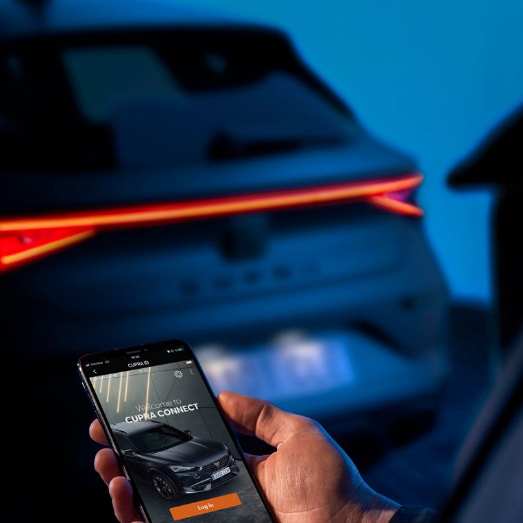 Osoba drží mobilný telefón, na ktorom je zobrazená aplikácia na ovládanie vozidla. V pozadí je vidieť zadnú časť tmavého auta s rozsvietenými svetlami. Prostredie je osvetlené modrým svetlom.