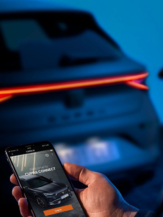 Osoba drží mobilný telefón, na ktorom je zobrazená aplikácia na ovládanie vozidla. V pozadí je vidieť zadnú časť tmavého auta s rozsvietenými svetlami. Prostredie je osvetlené modrým svetlom.