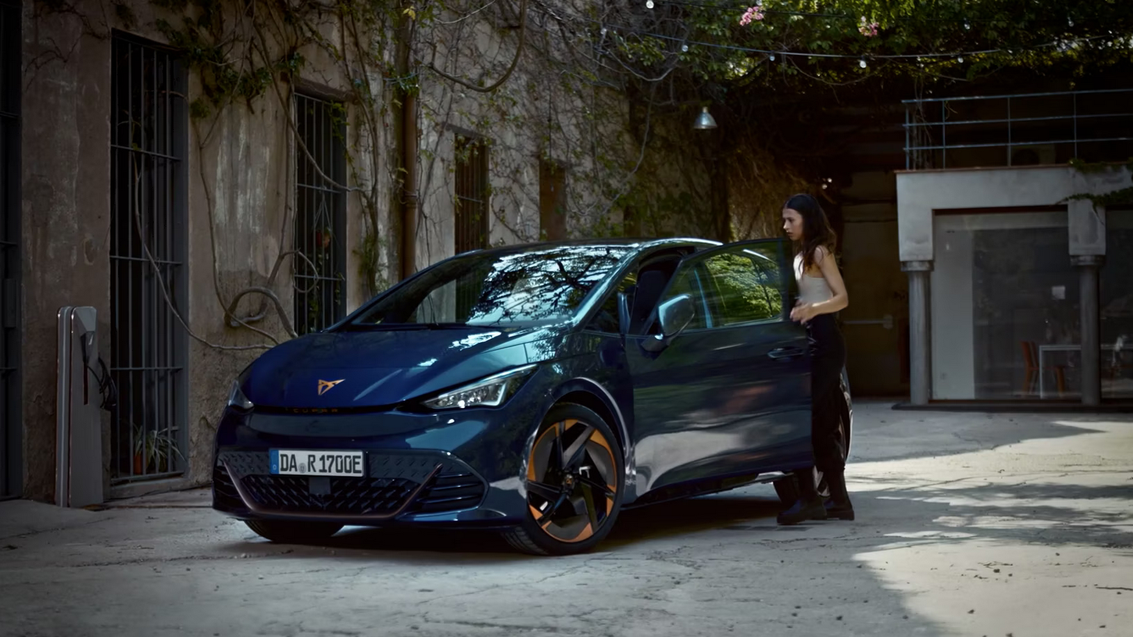 Žena stojí vedľa tmavomodrého športového auta, ktoré je zaparkované pred budovou s popínavými rastlinami. Vozidlo ma kolesá s elegantnými diskami.
