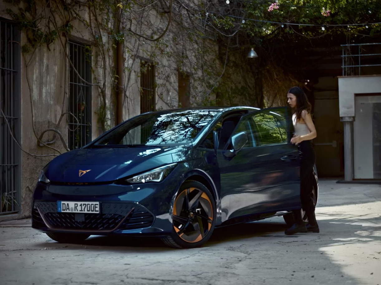 Žena stojí vedľa tmavomodrého športového auta, ktoré je zaparkované pred budovou s popínavými rastlinami. Vozidlo ma kolesá s elegantnými diskami.