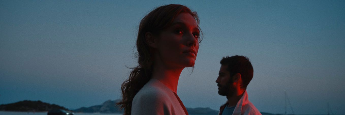 Žena a muž stoja pri pobreží za súmraku. V pozadí vidieť jachty na mori.