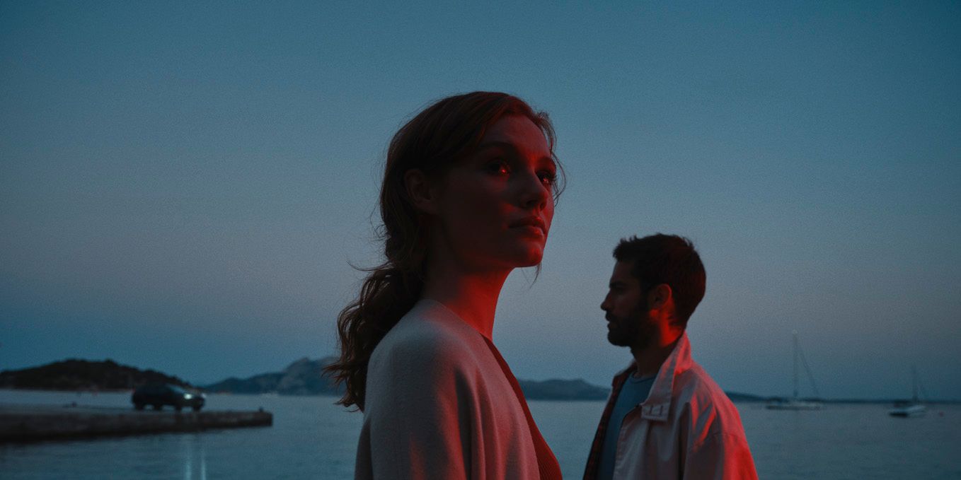 Žena a muž stoja pri pobreží za súmraku. V pozadí vidieť jachty na mori.