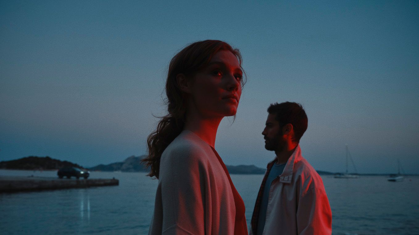 Žena a muž stoja pri pobreží za súmraku. V pozadí vidieť jachty na mori.