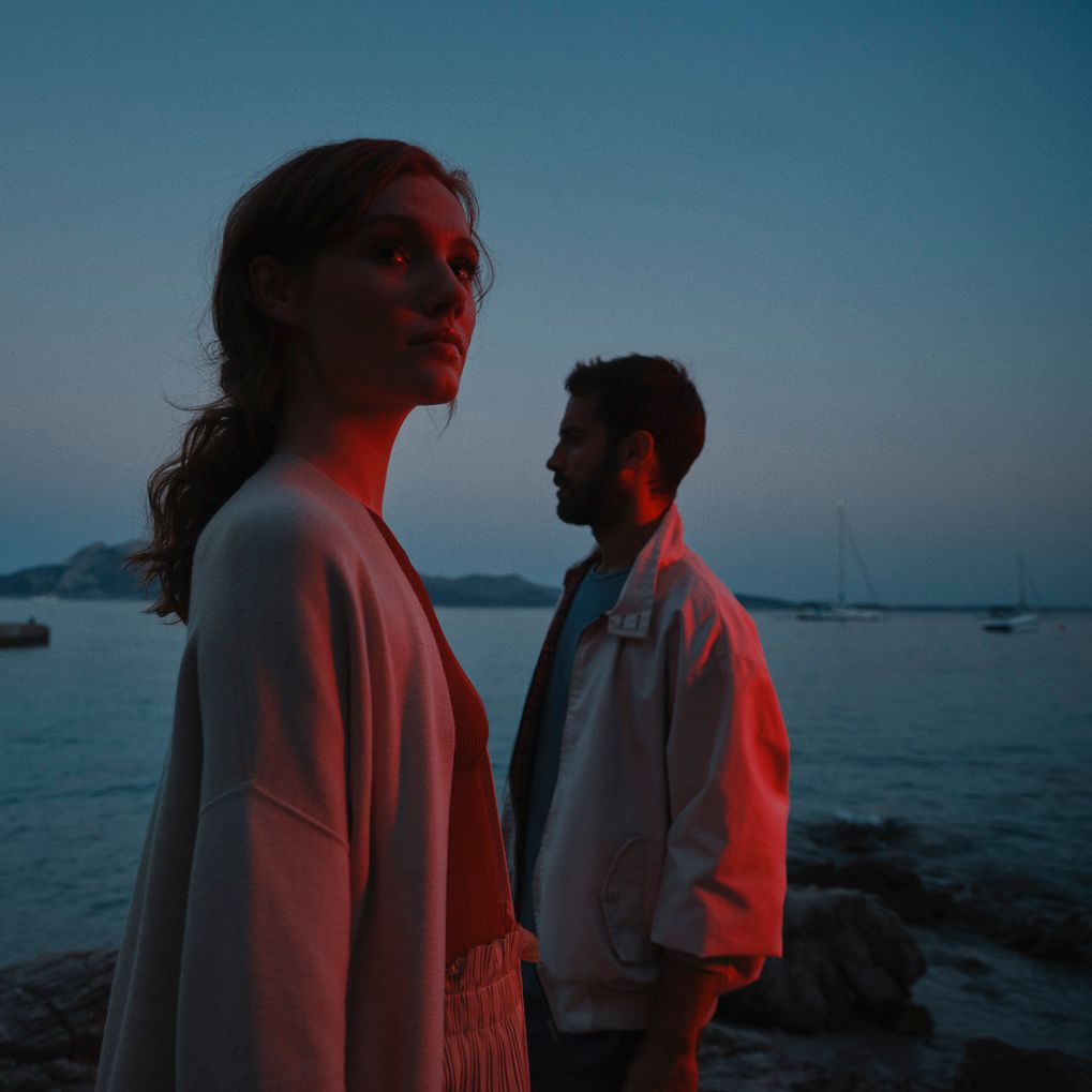 Žena a muž stoja pri pobreží za súmraku. V pozadí vidieť jachty na mori.