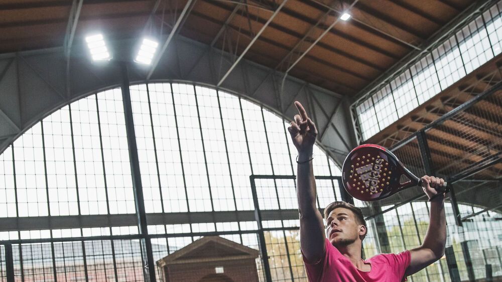 Muž hrá tenis na krytom padle kurte. Má zdvihnutú ruku a drží raketu. Pozadie tvorí veľké okno s kovovou konštrukciou.