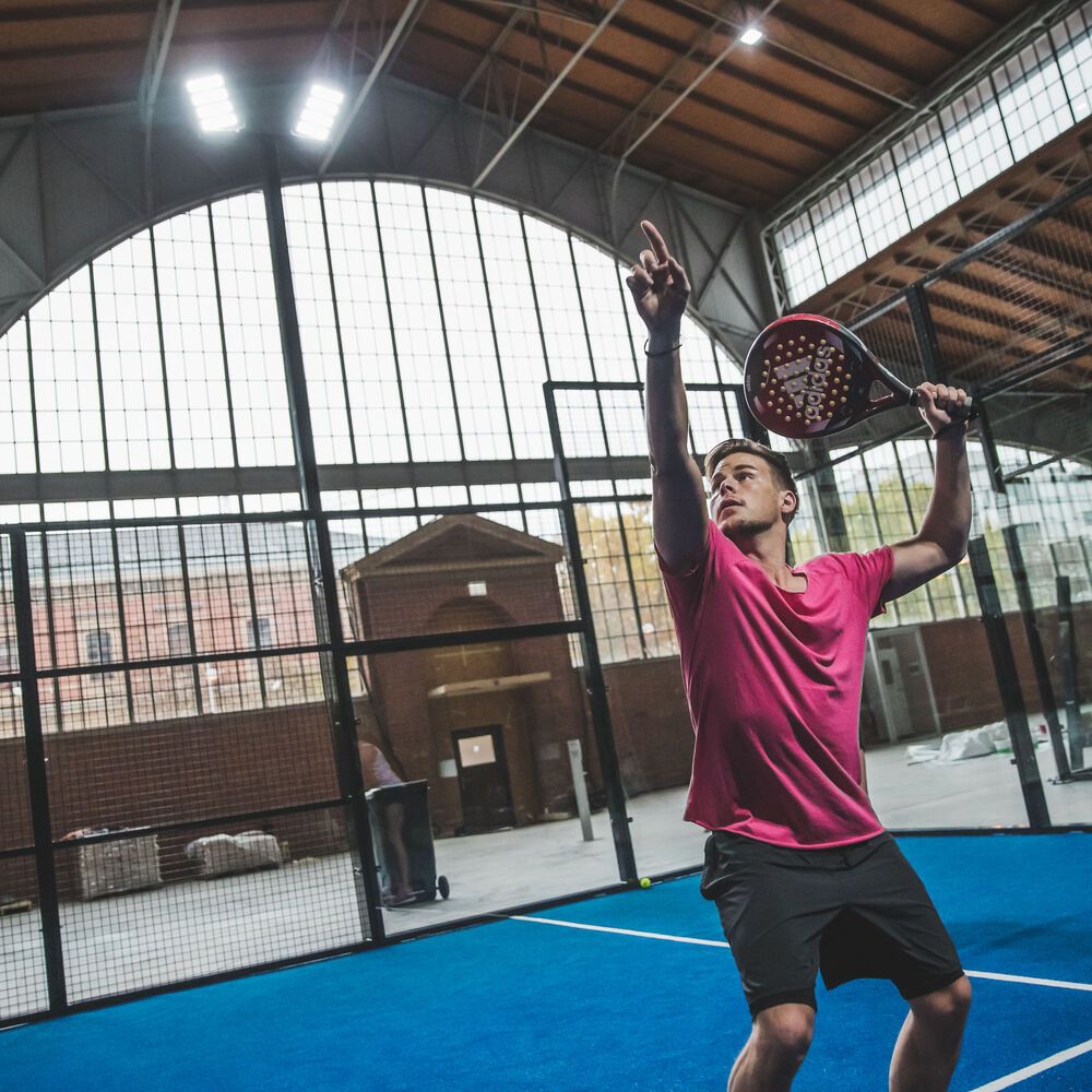 Muž hrá tenis na krytom padle kurte. Má zdvihnutú ruku a drží raketu. Pozadie tvorí veľké okno s kovovou konštrukciou.
