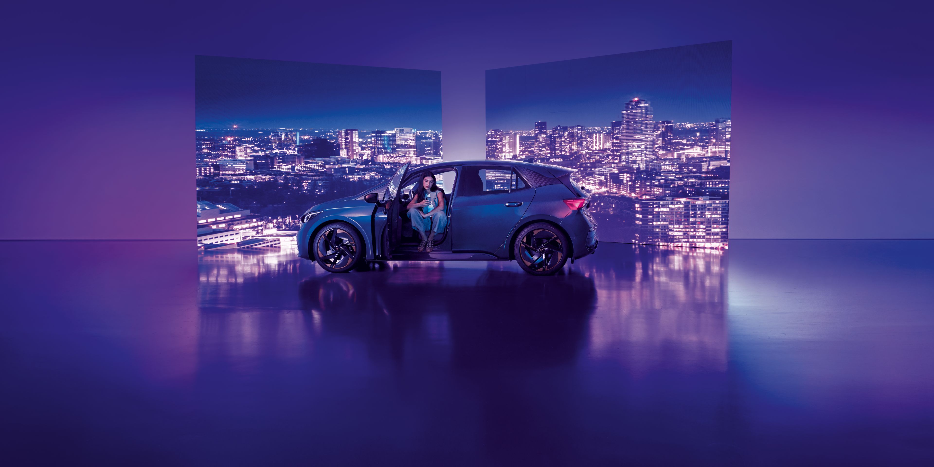 Pozadie tvoria dva veľké billboardy s panoramatickým výhľadom na nočné mesto, pred ktorými stojí moderné auto s otvorenými dverami. Žena sedí v aute na mieste vodiča, interiér je osvetlený jemným svetlom.