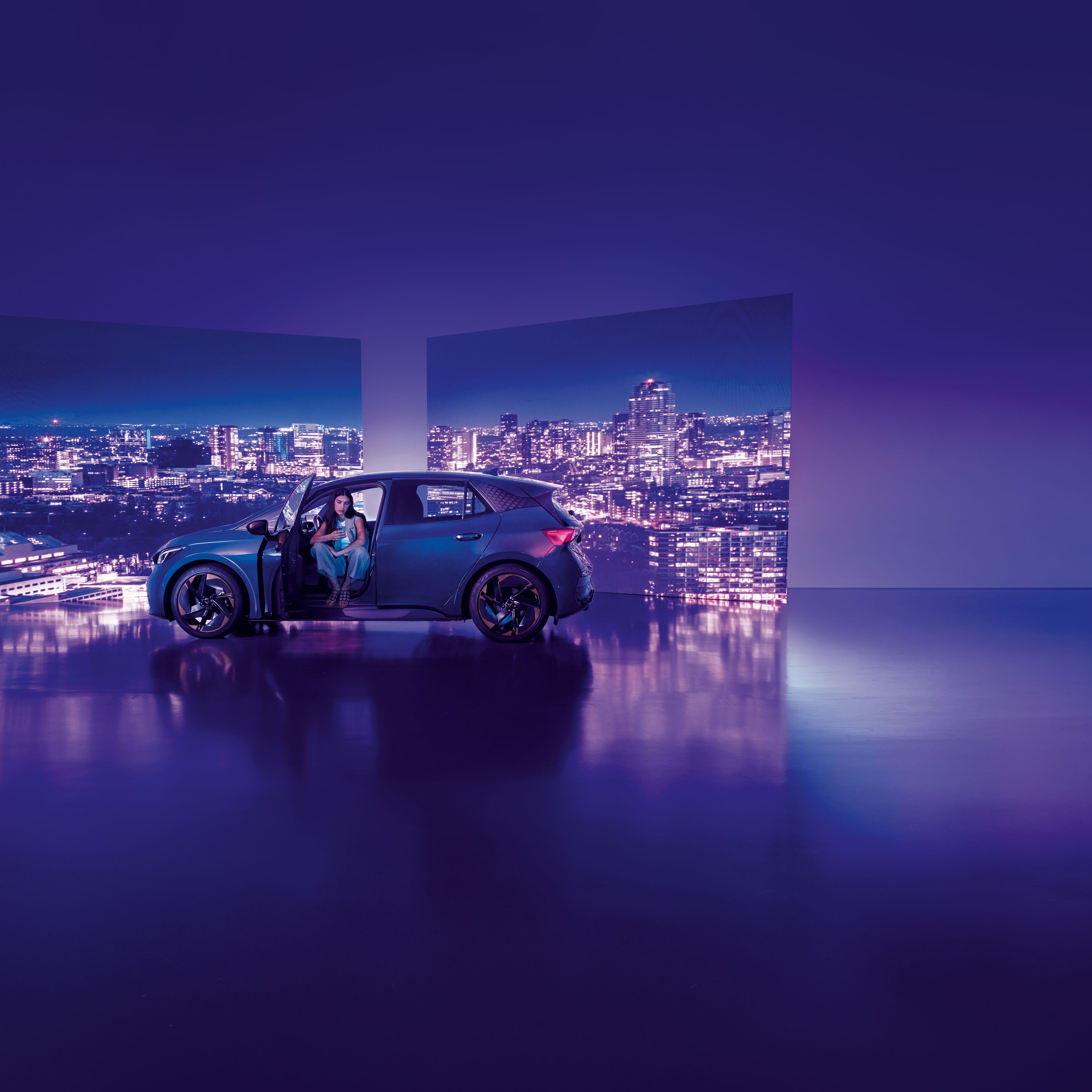 Pozadie tvoria dva veľké billboardy s panoramatickým výhľadom na nočné mesto, pred ktorými stojí moderné auto s otvorenými dverami. Žena sedí v aute na mieste vodiča, interiér je osvetlený jemným svetlom.