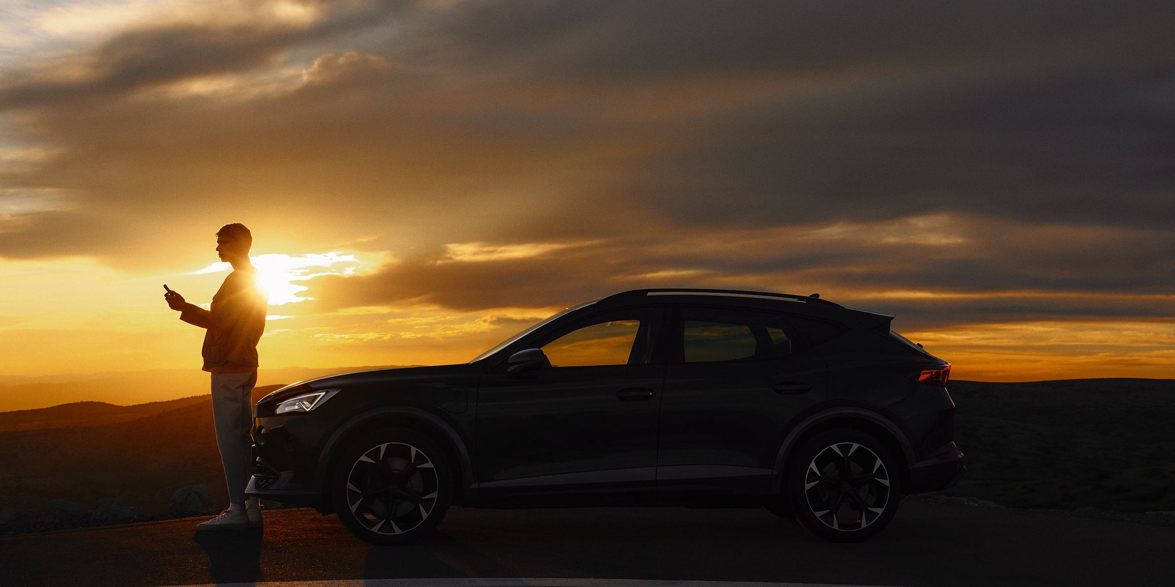 Osoba stojí vedľa zaparkovaného automobilu na kraji cesty pri západe slnka. Obloha je pokrytá tmavými mrakmi, ktoré vytvárajú dramatický efekt svetla. 