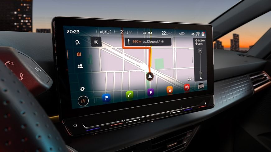 Auto s moderným dotykovým displejom s navigáciou. Na okraji obrazovky sa nachádzajú ovládacie prvky klimatizácie a ďalších funkcií vozidla. V pozadí je vidieť mesto pri západe slnka.