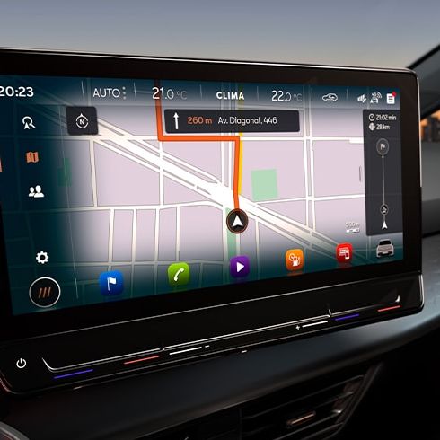 Auto s moderným dotykovým displejom s navigáciou. Na okraji obrazovky sa nachádzajú ovládacie prvky klimatizácie a ďalších funkcií vozidla. V pozadí je vidieť mesto pri západe slnka.