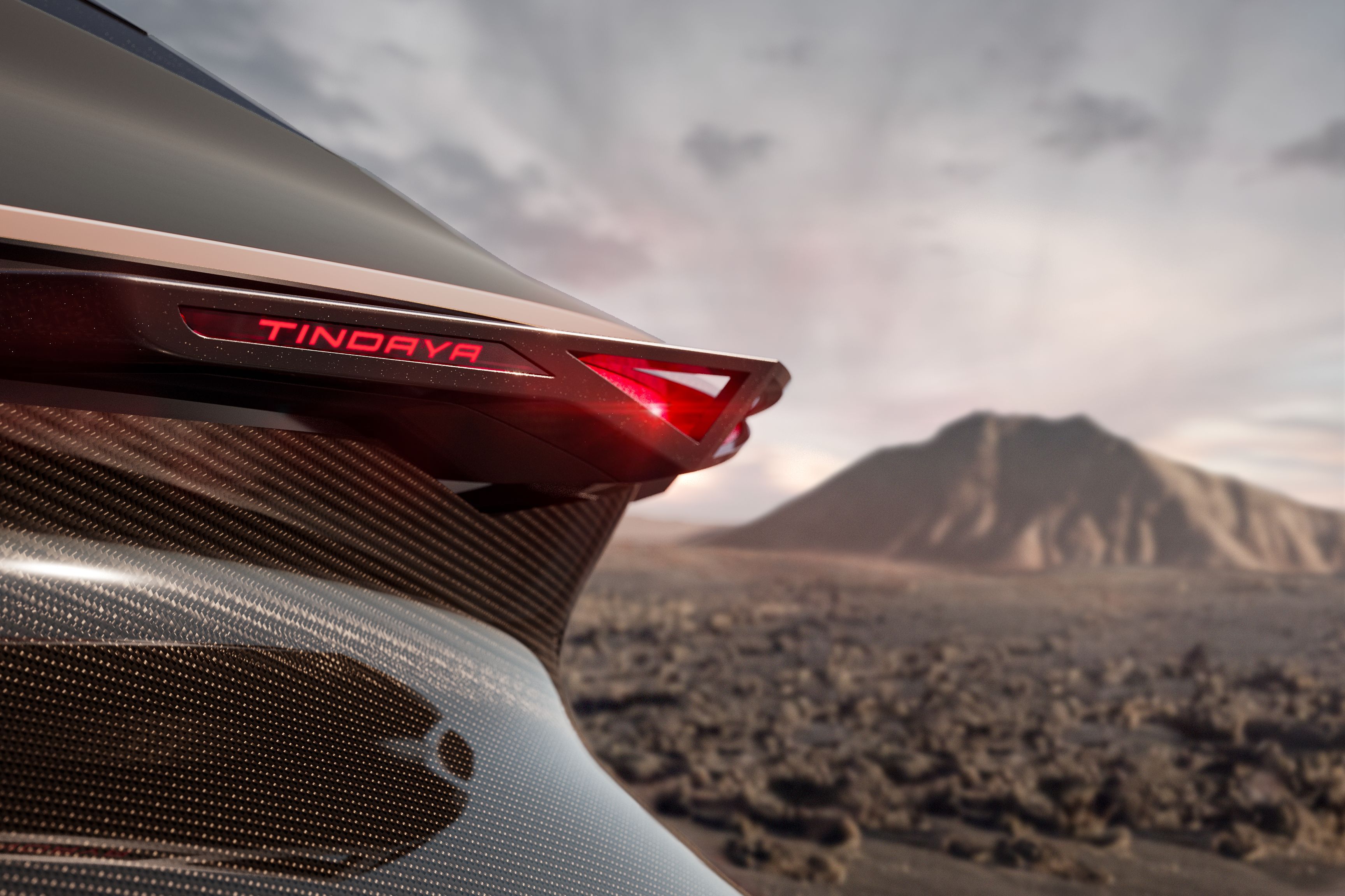 Detailný záber na zadné svetlo futuristického automobilu s názvom 'TINDAYA'. V pozadí je krajinná scenéria, pravdepodobne púšť alebo kamenistá planina. Svetlo svieti červenou farbou.