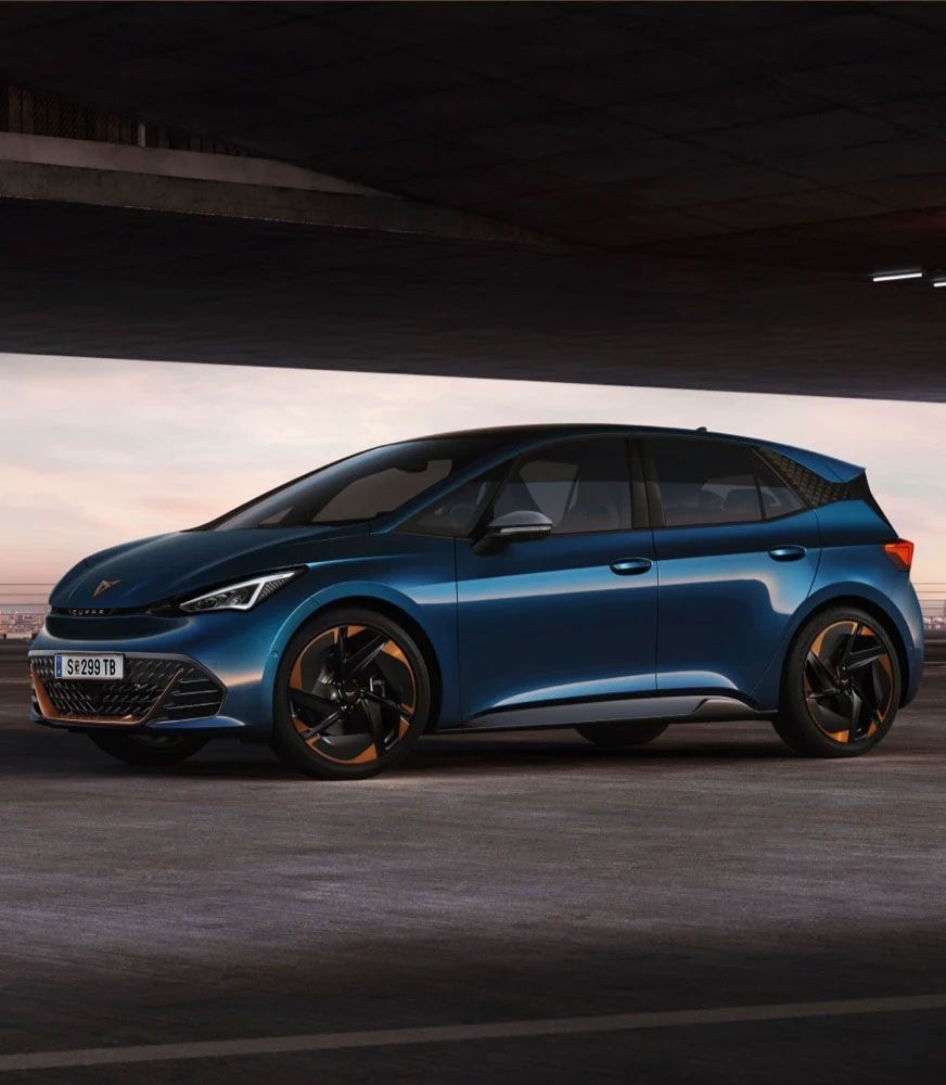 Tmavomodrý CUPRA Born, zaparkovaný pod betónovým mostom, s čiernymi diskami s bronzovými prvkami. Na auto dopadajú jemné lúče svetla, ktoré zvýrazňujú jeho moderný dizajn. 