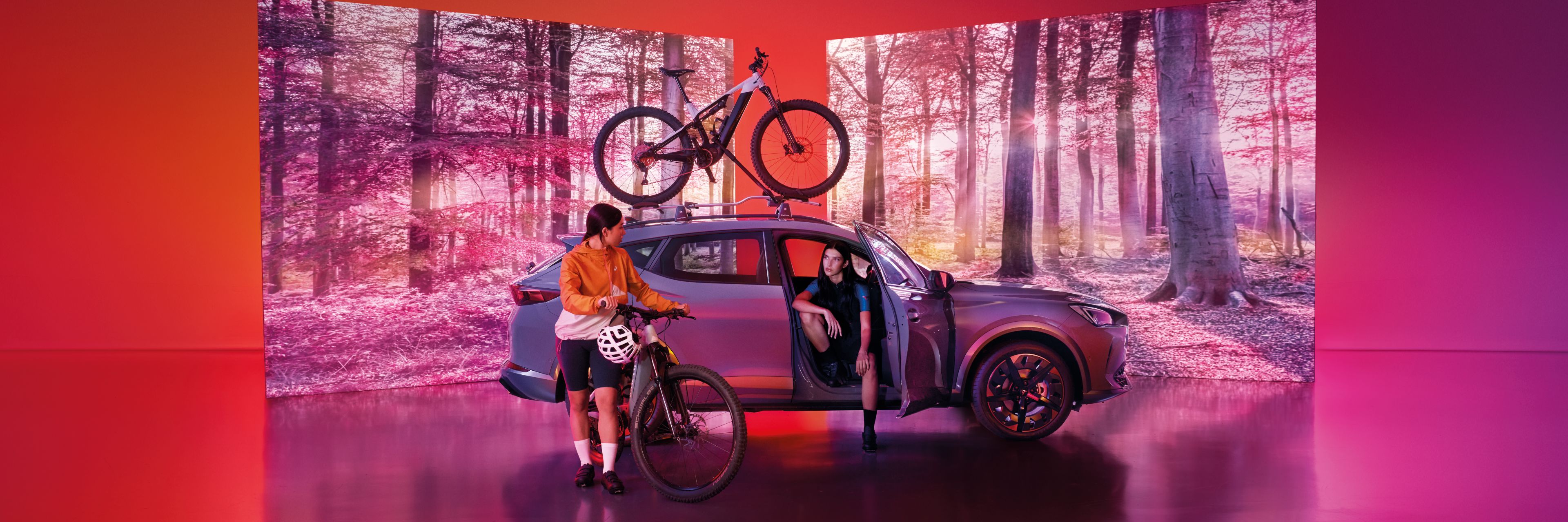 Vozidlo CUPRA so strešným nosičom s upevneným vozidlom stojí pred dvomi obrazovkami, na ktorých je les. Žena sedí vo vozidle. Druhá žena s bicyklom stojí pri vozidle.