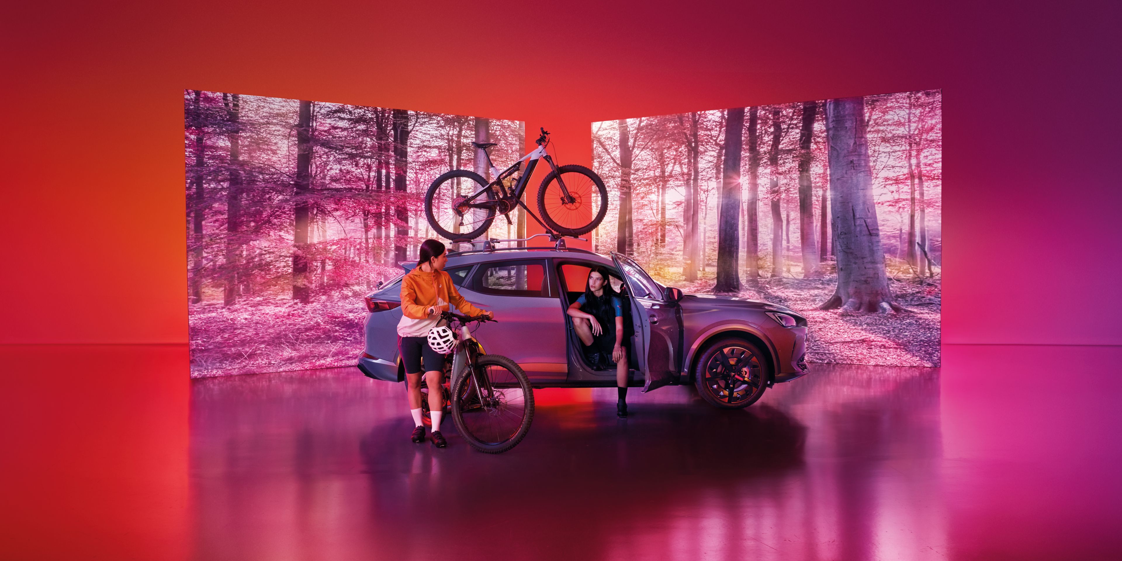 Vozidlo CUPRA so strešným nosičom s upevneným vozidlom stojí pred dvomi obrazovkami, na ktorých je les. Žena sedí vo vozidle. Druhá žena s bicyklom stojí pri vozidle.