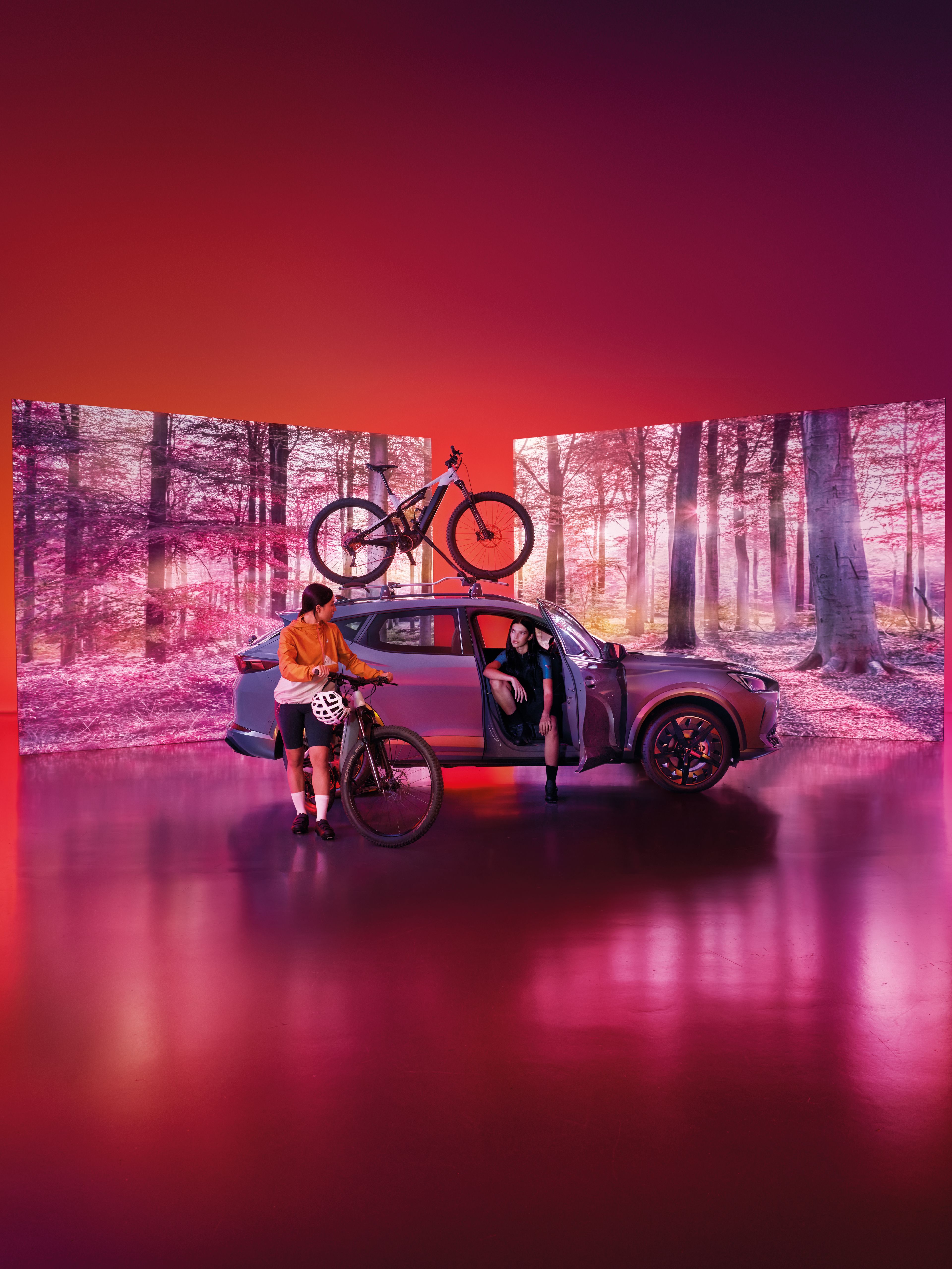 Vozidlo CUPRA so strešným nosičom s upevneným vozidlom stojí pred dvomi obrazovkami, na ktorých je les. Žena sedí vo vozidle. Druhá žena s bicyklom stojí pri vozidle.