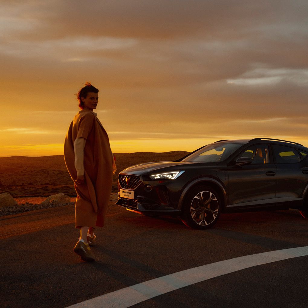 Žena prichádza k modelu CUPRA Formentor, ktorý stojí na ceste s výhľadom do údolia a na kopce, celú scénu osvetľuje zapadajúce slnko.
