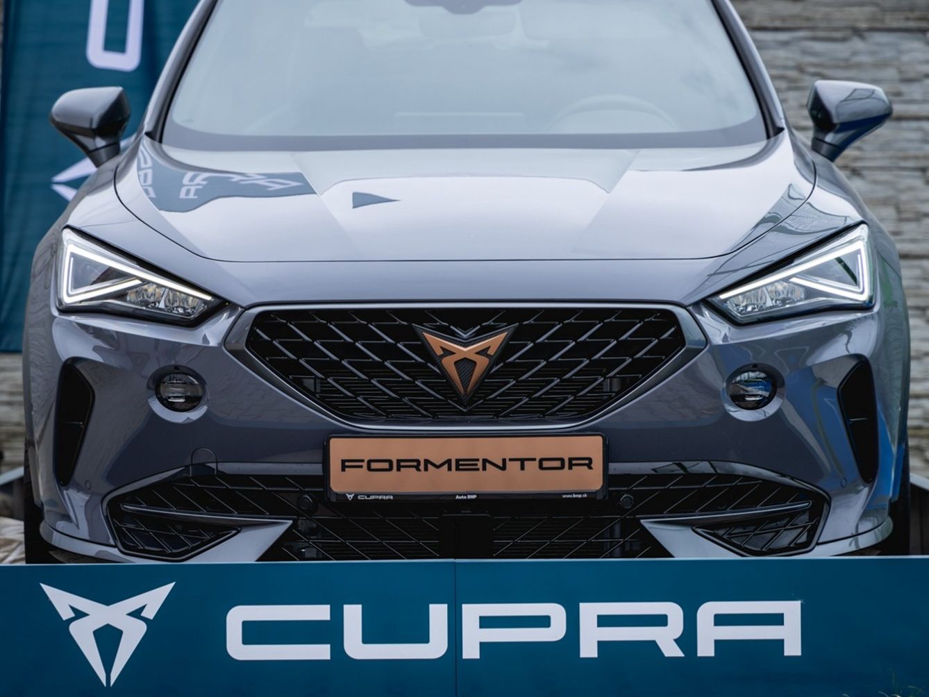 Predná časť tmavomodrého vozidla CUPRA Formentor. Sú viditeľné jeho charakteristické predné svetlá, maska chladiča a logo. Na spodnej časti je značka a logo.