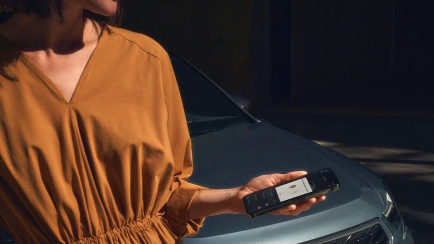 Žena v šatách drží v ruke smartfón pri modrom vozidle značky CUPRA.