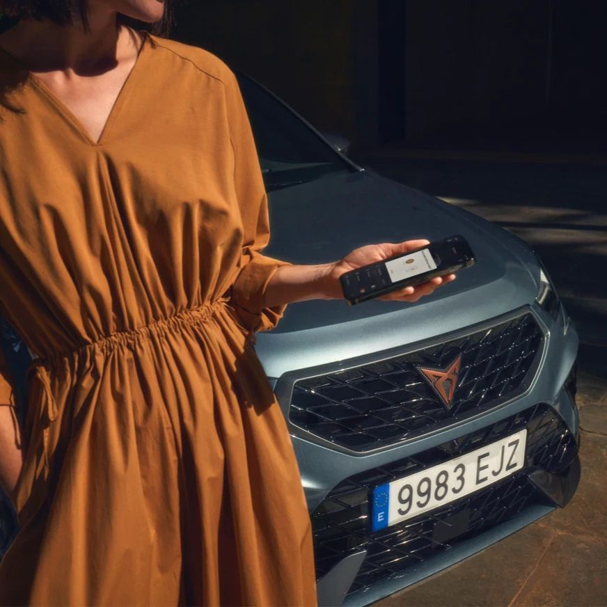 Žena v šatách drží v ruke smartfón pri modrom vozidle značky CUPRA.