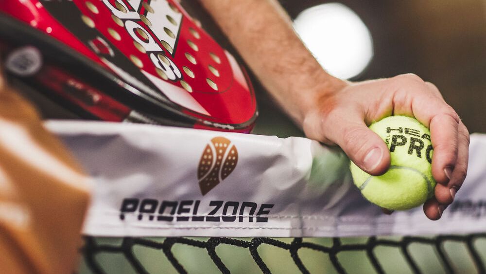 Osoba drží loptičku a raketu na padel, stojí blízko siete s logom "PADELZONE".