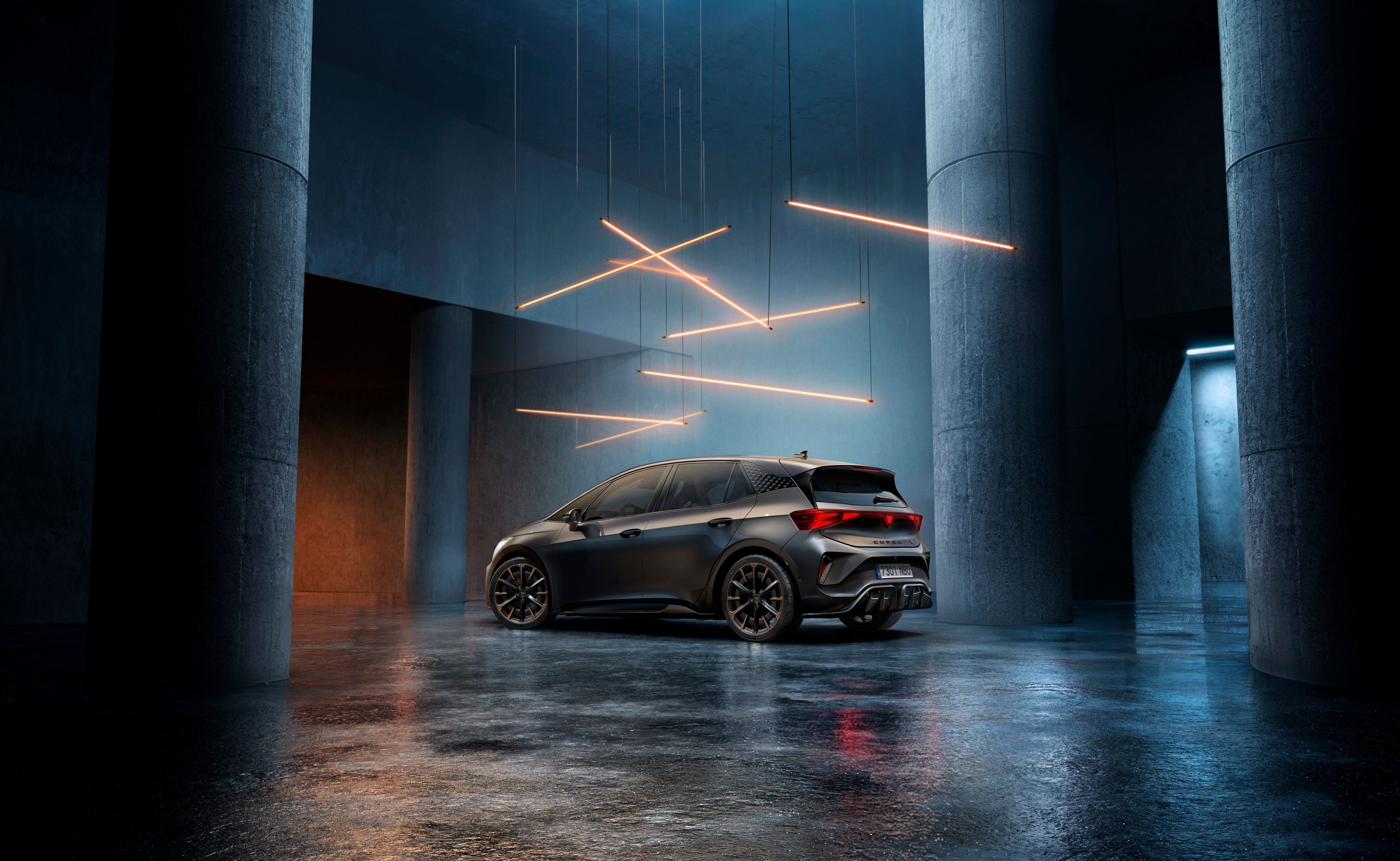 CUPRA Born stojí v štýlovo osvetlenom priemyselnom priestore s betónovými stenami a stĺpmi. Zo stropu visí niekoľko lineárnych svetelných zdrojov, ktoré vytvárajú elegantnú atmosféru. Vozidlo je vidieť zo zadnej strany a odráža svetlo na lesklej podlahe.