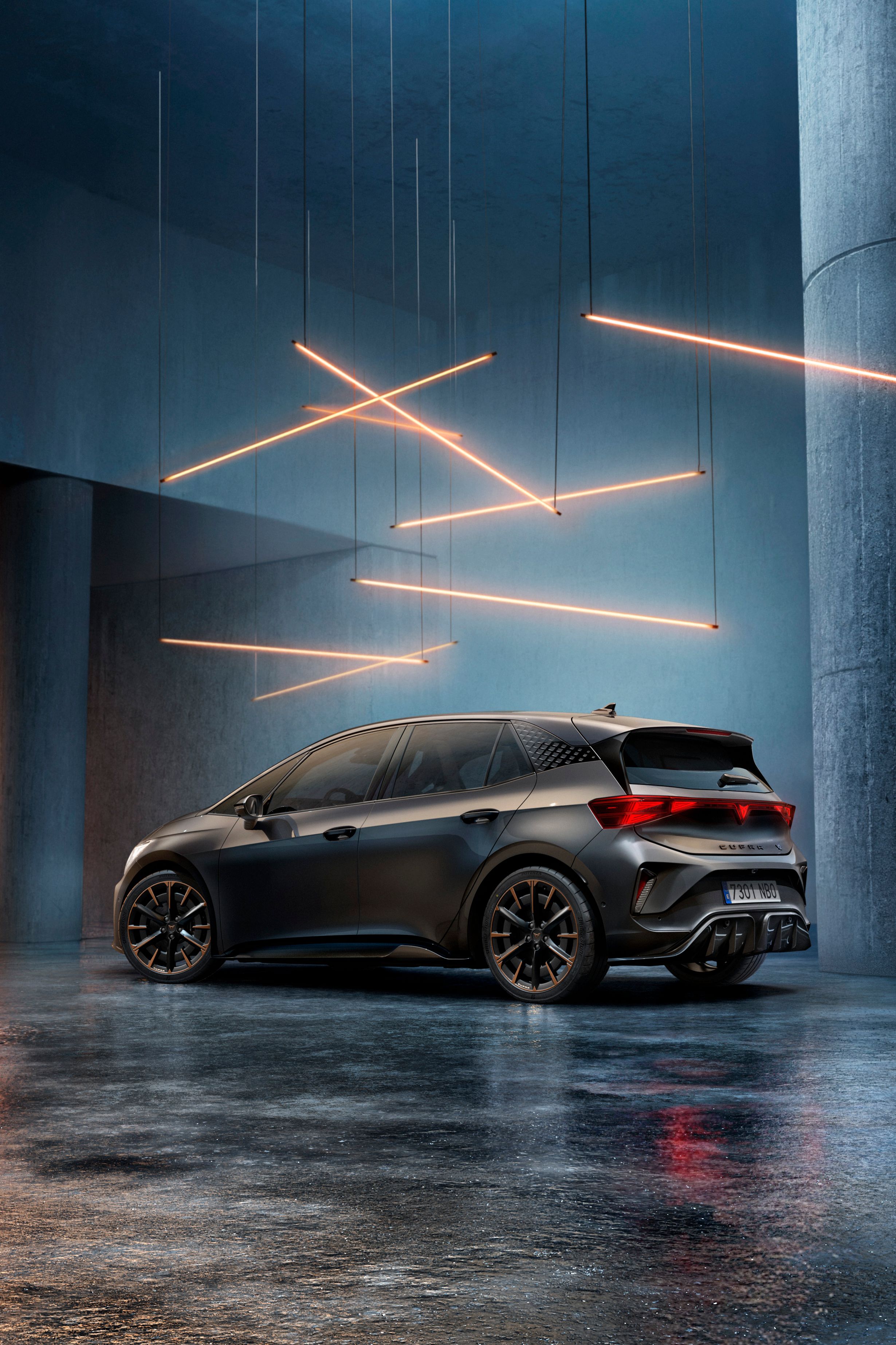 CUPRA Born stojí v štýlovo osvetlenom priemyselnom priestore s betónovými stenami a stĺpmi. Zo stropu visí niekoľko lineárnych svetelných zdrojov, ktoré vytvárajú elegantnú atmosféru. Vozidlo je vidieť zo zadnej strany a odráža svetlo na lesklej podlahe.