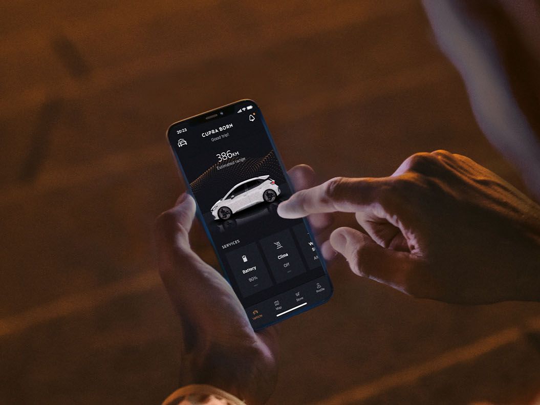 Človek ovláda nastavenia auta pomocou mobilnej aplikácie na smartfóne. Tmavé pozadie a osvetlenie naznačujú večer. Muž ukazuje prstom na obrazovku, kde je zobrazené auto.