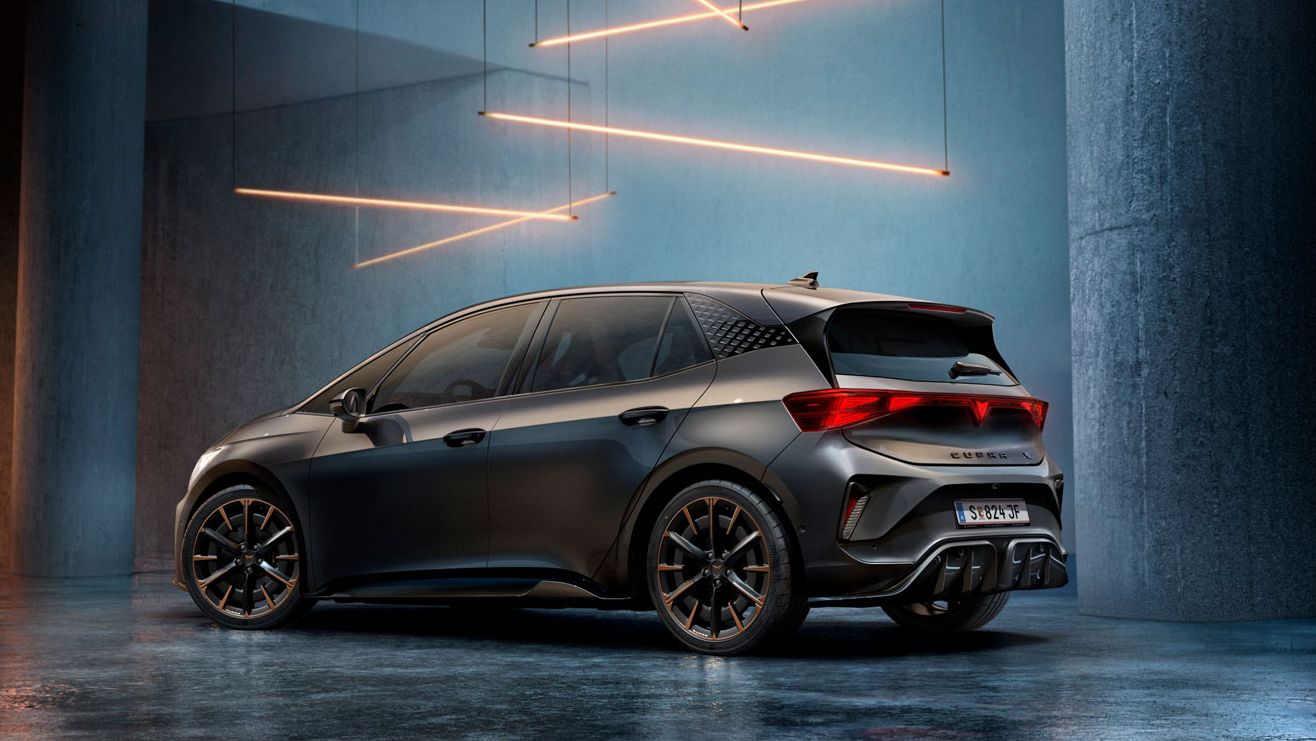 CUPRA Born stojí v štýlovo osvetlenom priemyselnom priestore s betónovými stenami a stĺpmi. Zo stropu visí niekoľko lineárnych svetelných zdrojov, ktoré vytvárajú elegantnú atmosféru. Vozidlo je vidieť zo zadnej strany a odráža svetlo na lesklej podlahe.