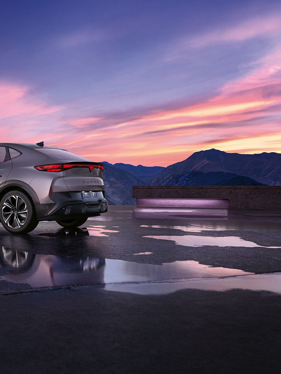 Moderné SUV so šedou metalízou je zaparkované na mokrej ploche s horami v pozadí. Obloha je sfarbená do ružova pri západe slnka. Auto má elegantný dizajn s aerodynamickými líniami.