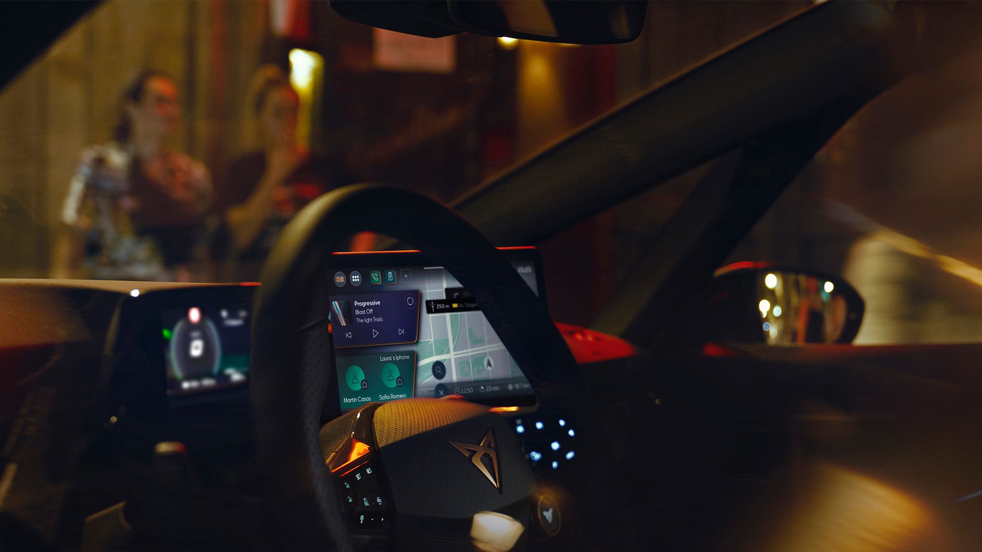 Interiér automobilu s dobre viditeľným dotykovým displejom navigácie a ovládacími prvkami na volante. V navigácii sú zobrazené informácie o trase. Na pozadí je rozmazaný vonkajší pohľad na ulicu.