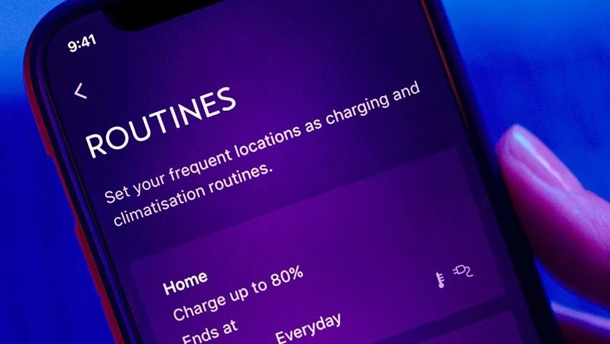 Mobilný telefón zobrazuje aplikáciu na nastavenie rutín nabíjania a klimatizácie. Na displeji sú dve prednastavené rutiny pre domov a prácu. Pozadie je modré.
