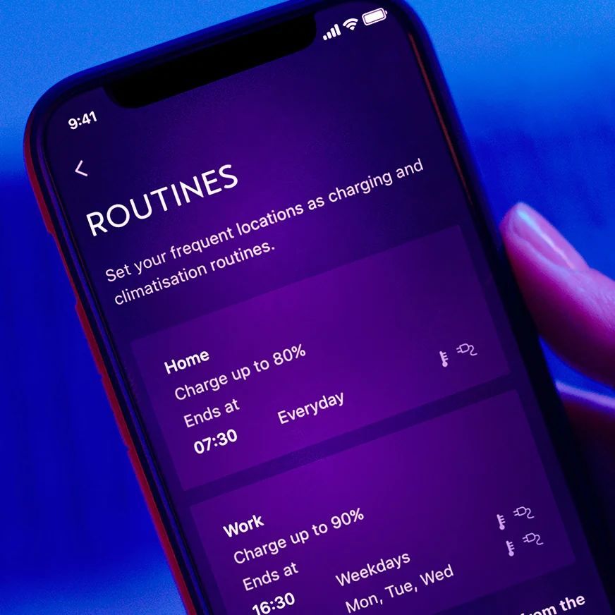 Mobilný telefón zobrazuje aplikáciu na nastavenie rutín nabíjania a klimatizácie. Na displeji sú dve prednastavené rutiny pre domov a prácu. Pozadie je modré.