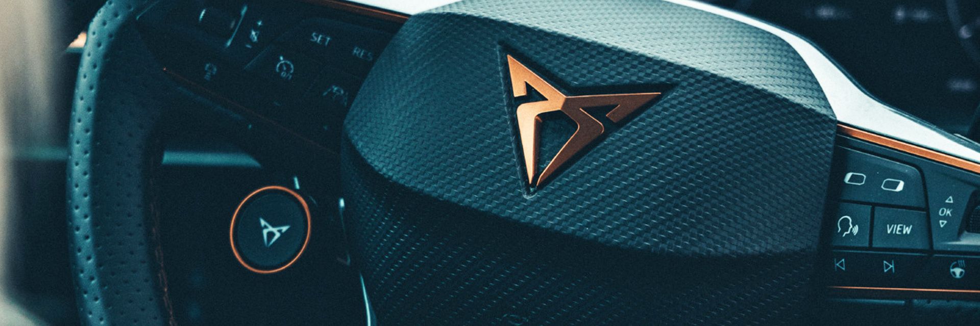 Detailný záber na volant moderného auta značky CUPRA s výrazným logom v strede. Na volante sú rôzne tlačidlá a ovládače. V pozadí je prístrojový panel.