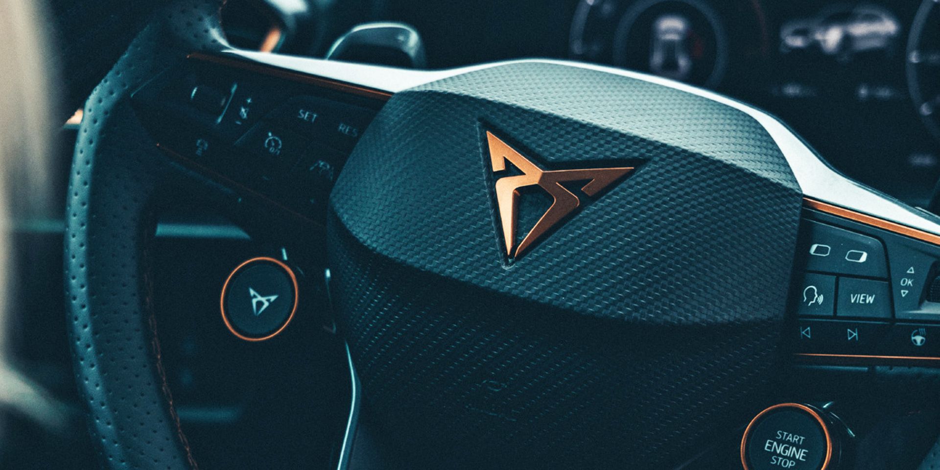 Detailný záber na volant moderného auta značky CUPRA s výrazným logom v strede. Na volante sú rôzne tlačidlá a ovládače. V pozadí je prístrojový panel.