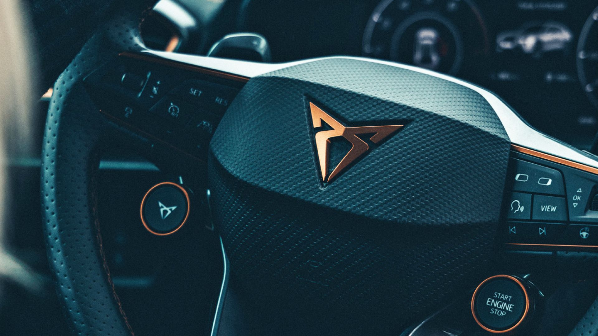 Detailný záber na volant moderného auta značky CUPRA s výrazným logom v strede. Na volante sú rôzne tlačidlá a ovládače. V pozadí je prístrojový panel.