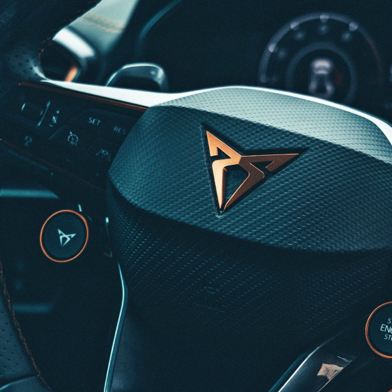 Detailný záber na volant moderného auta značky CUPRA s výrazným logom v strede. Na volante sú rôzne tlačidlá a ovládače. V pozadí je prístrojový panel.