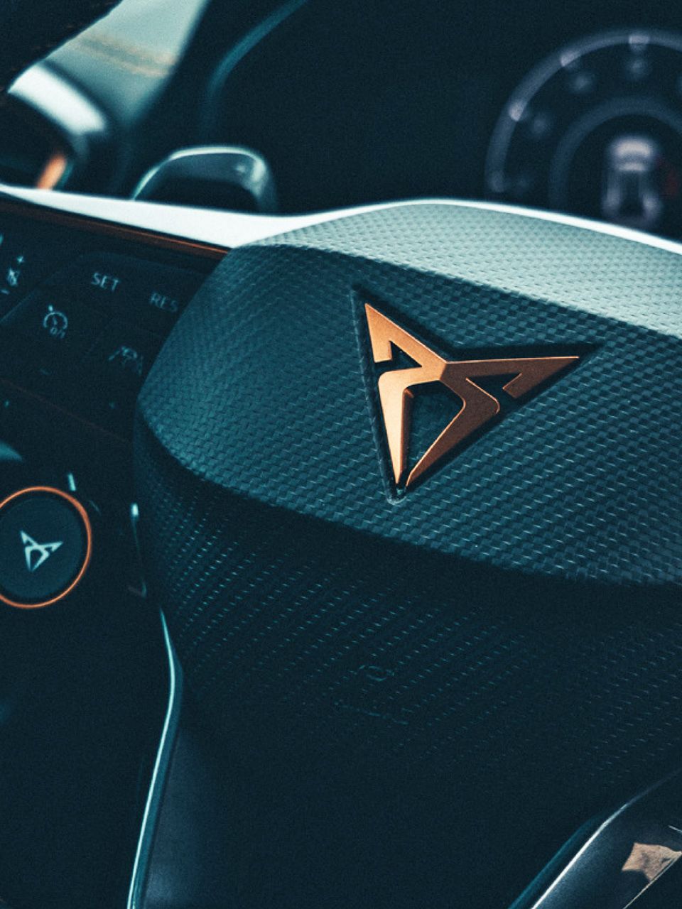 Detailný záber na volant moderného auta značky CUPRA s výrazným logom v strede. Na volante sú rôzne tlačidlá a ovládače. V pozadí je prístrojový panel.