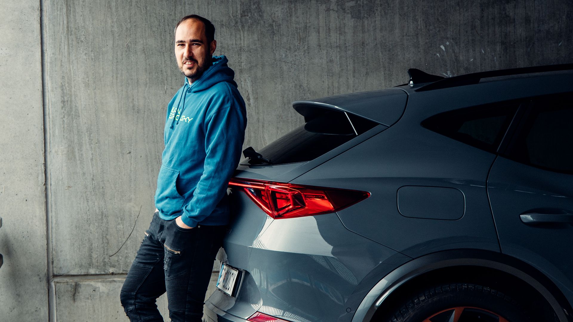 Muž v modrej mikine sa opiera o tmavosivé auto na parkovisku. Auto má moderný dizajn s výraznými zadnými svetlami. Pozadie tvorí betónová stena.