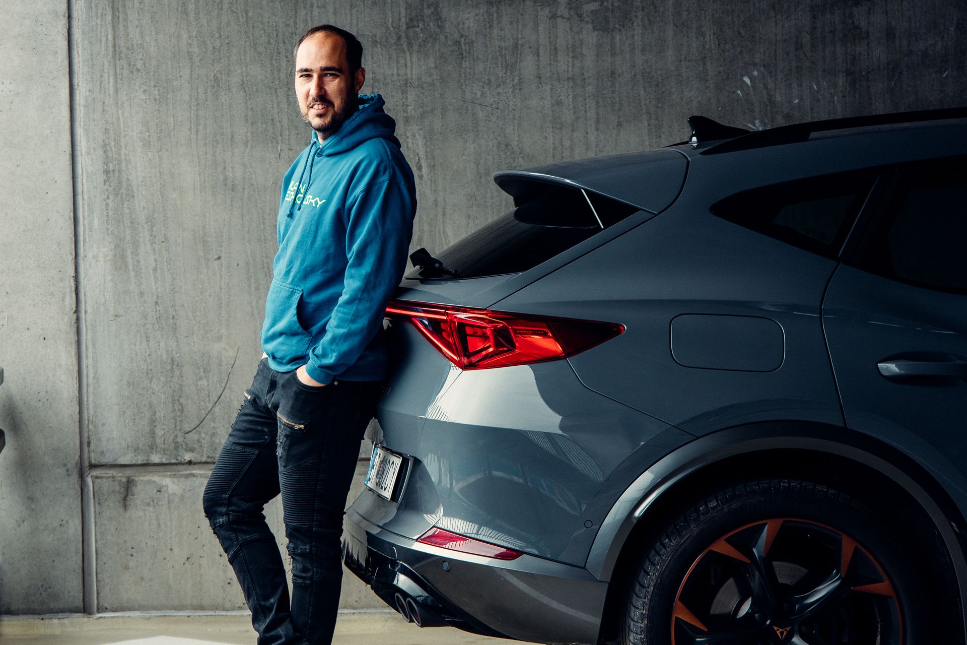 Muž v modrej mikine sa opiera o tmavosivé auto na parkovisku. Auto má moderný dizajn s výraznými zadnými svetlami. Pozadie tvorí betónová stena.