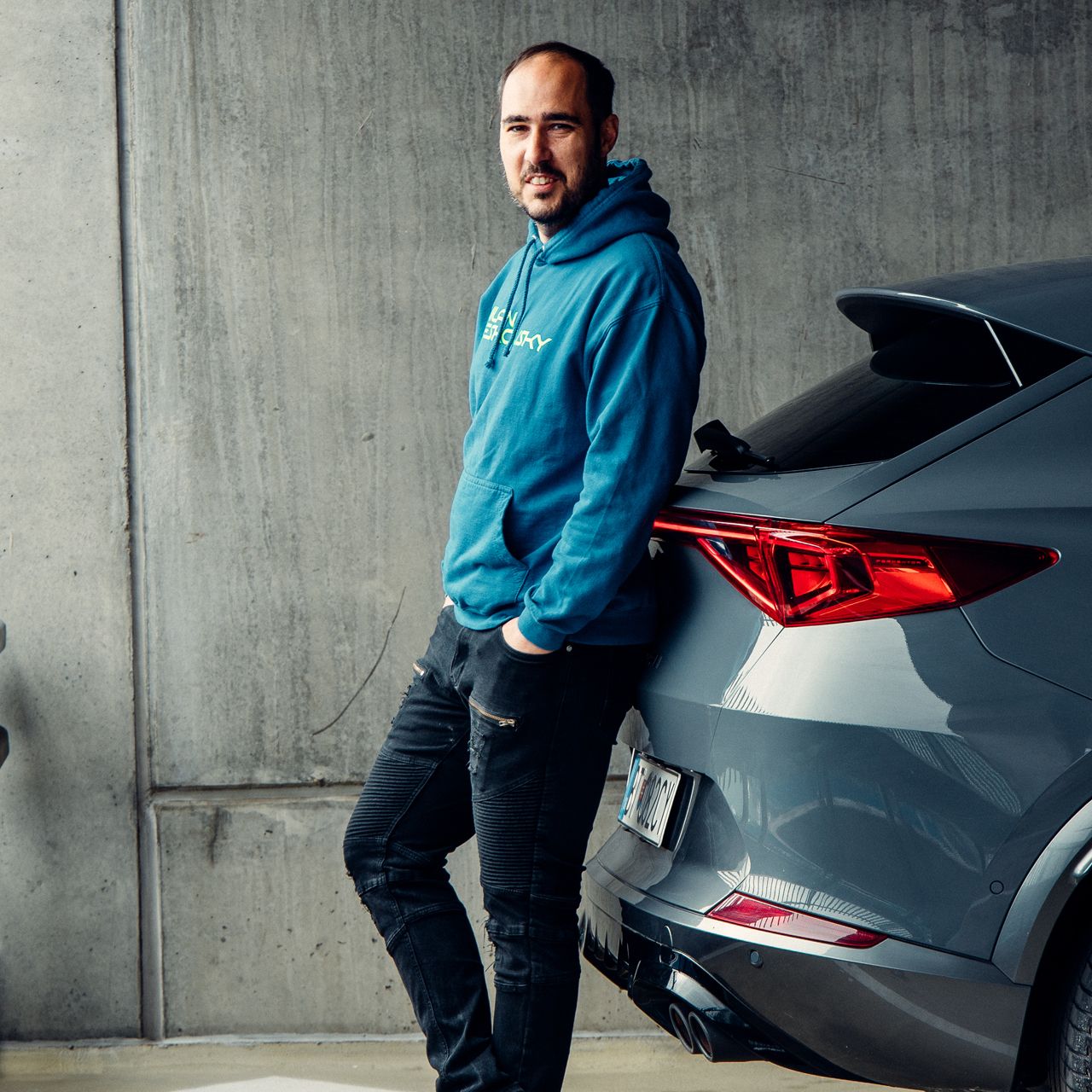 Muž v modrej mikine sa opiera o tmavosivé auto na parkovisku. Auto má moderný dizajn s výraznými zadnými svetlami. Pozadie tvorí betónová stena.