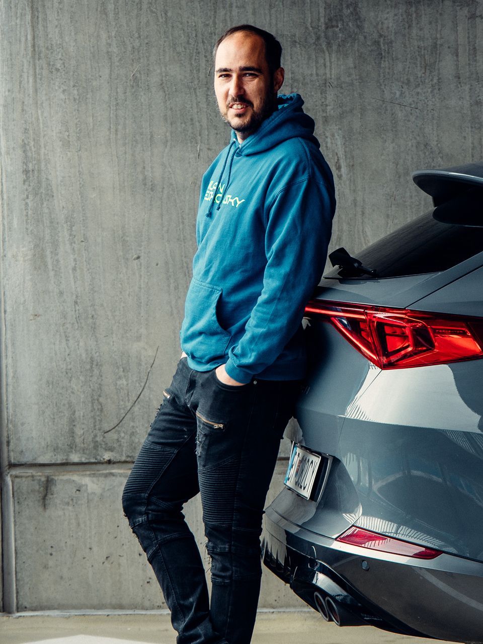 Muž v modrej mikine sa opiera o tmavosivé auto na parkovisku. Auto má moderný dizajn s výraznými zadnými svetlami. Pozadie tvorí betónová stena.