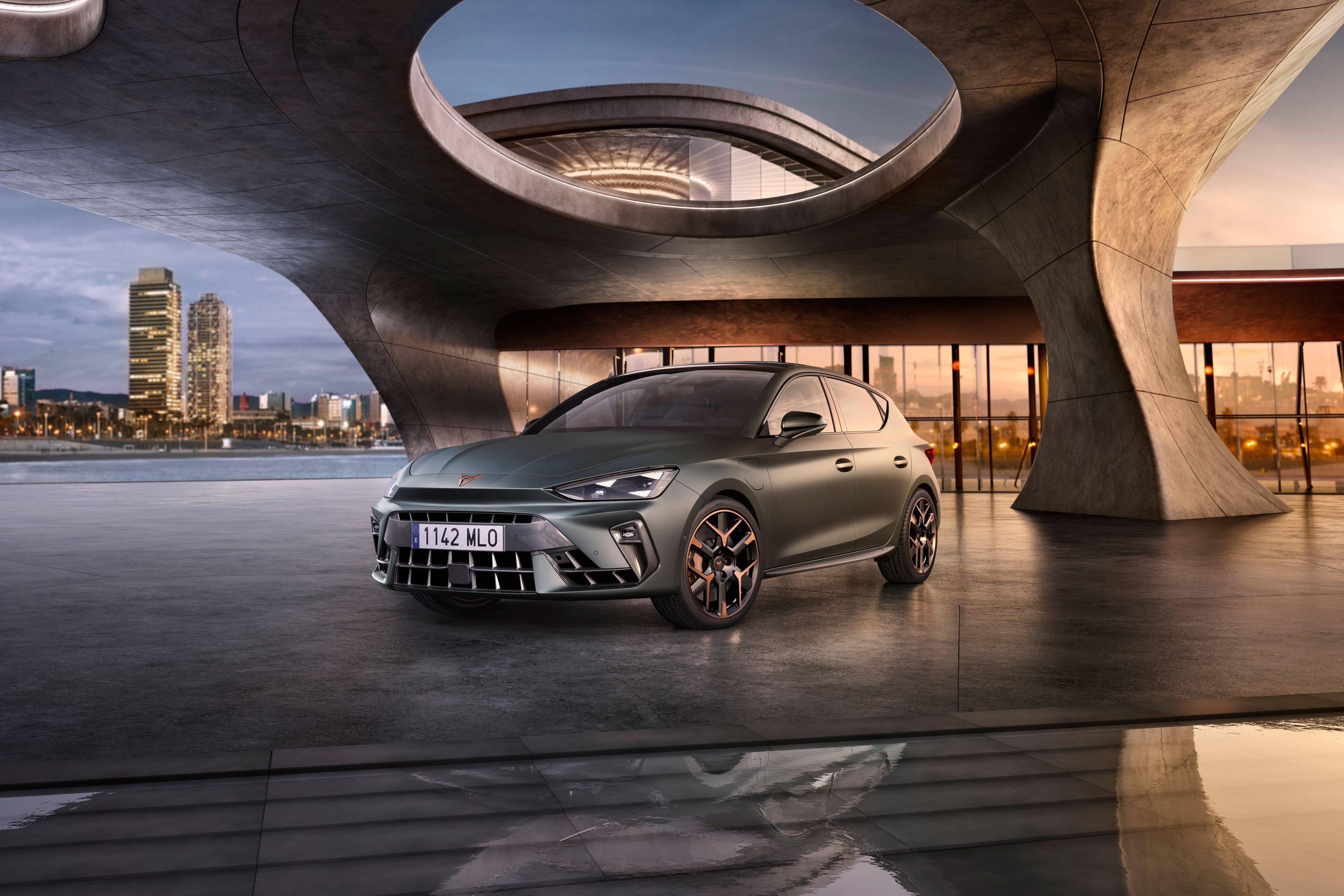 Automobil stojí na móle s modernou architektúrou v pozadí. Okolo je voda, na ktorej sa odráža okolie. V diaľke je silueta mesta.