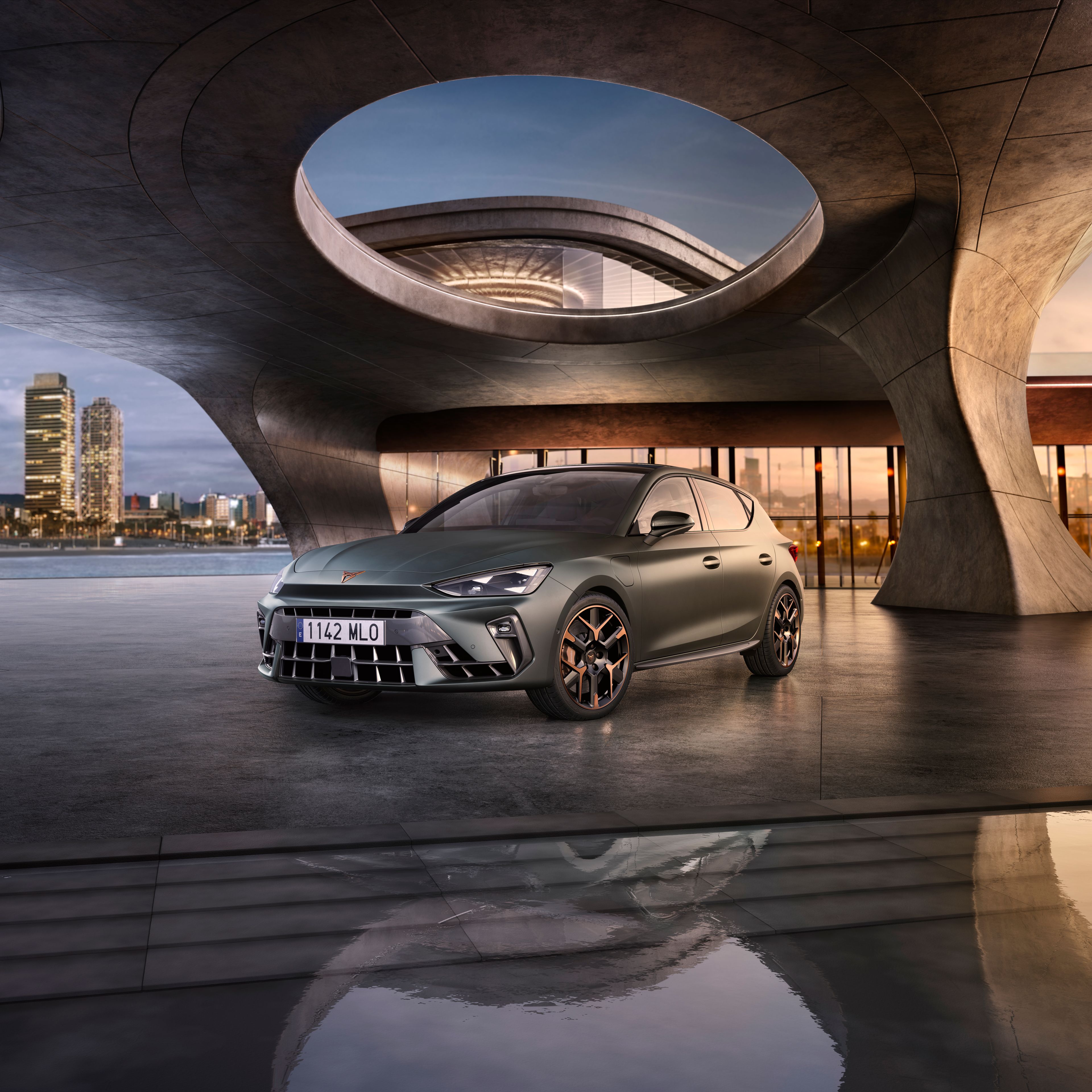 Automobil stojí na móle s modernou architektúrou v pozadí. Okolo je voda, na ktorej sa odráža okolie. V diaľke je silueta mesta.