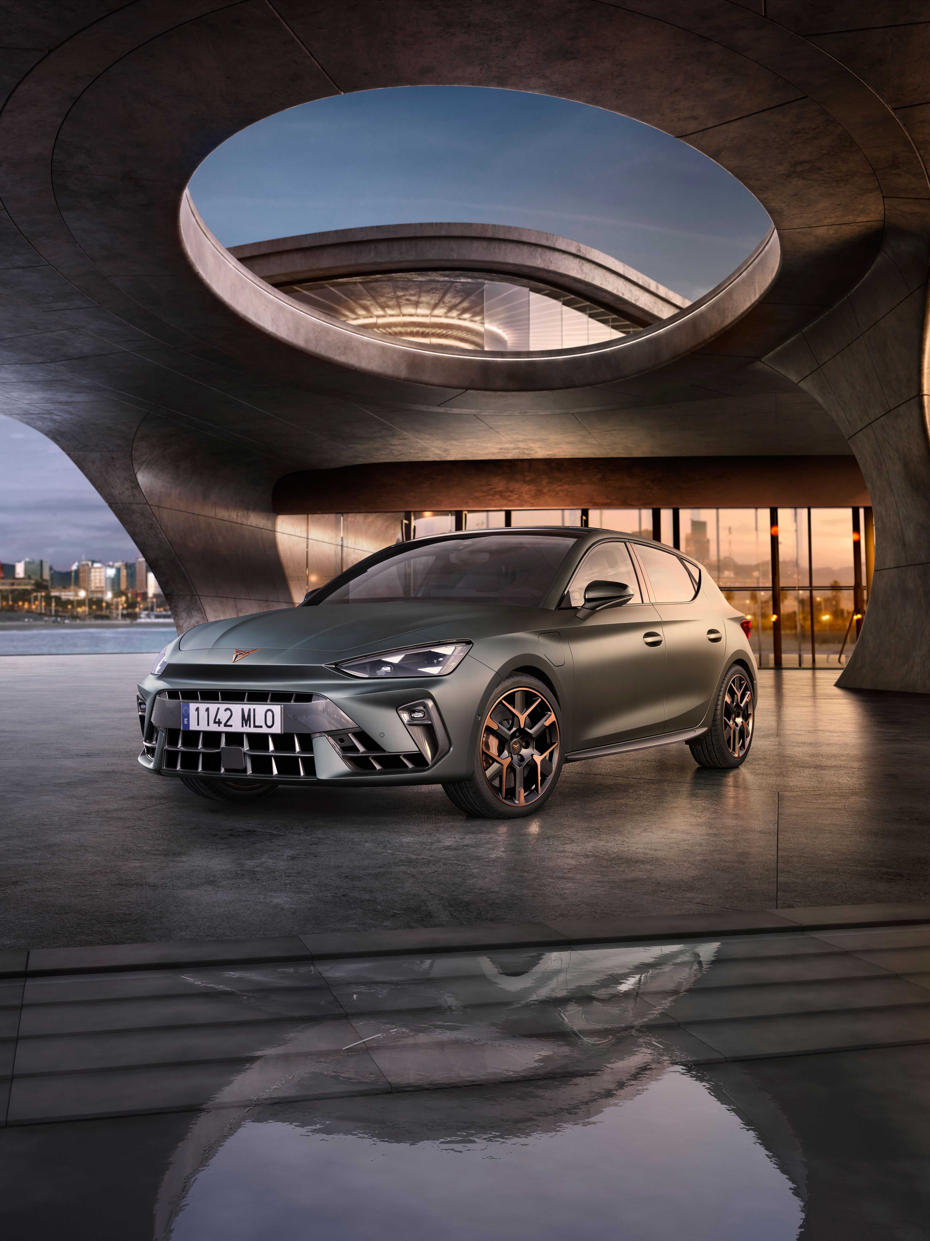 Automobil stojí na móle s modernou architektúrou v pozadí. Okolo je voda, na ktorej sa odráža okolie. V diaľke je silueta mesta.